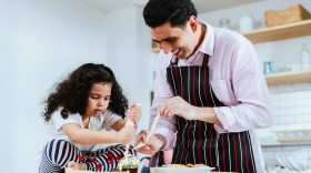 Father, fully involved in the process, guides his child's small hands as she intricately decorate cupcakes with cream, sprinkles, and edible delights.